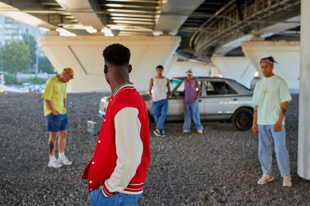 A diverse group of young friends hanging out under a city bridge with a car in the background.